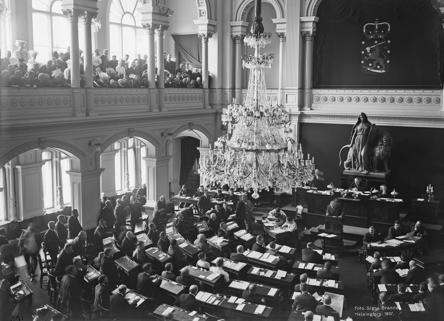 Valokuvassa valtiopäivien istunto Palokunnantalon juhlasalissa. Yleisöä kuuntelemassa istuntoa lehterillä. Kuvan keskellä näyttävä kattokruunu. Salin etuosaa koristaa Walter Runebergin Lex-veistoksen kipsikopio, jossa on miekkaa ja kilpeä pitelevä oikeuden jumalatar, jonka takana on leijona.