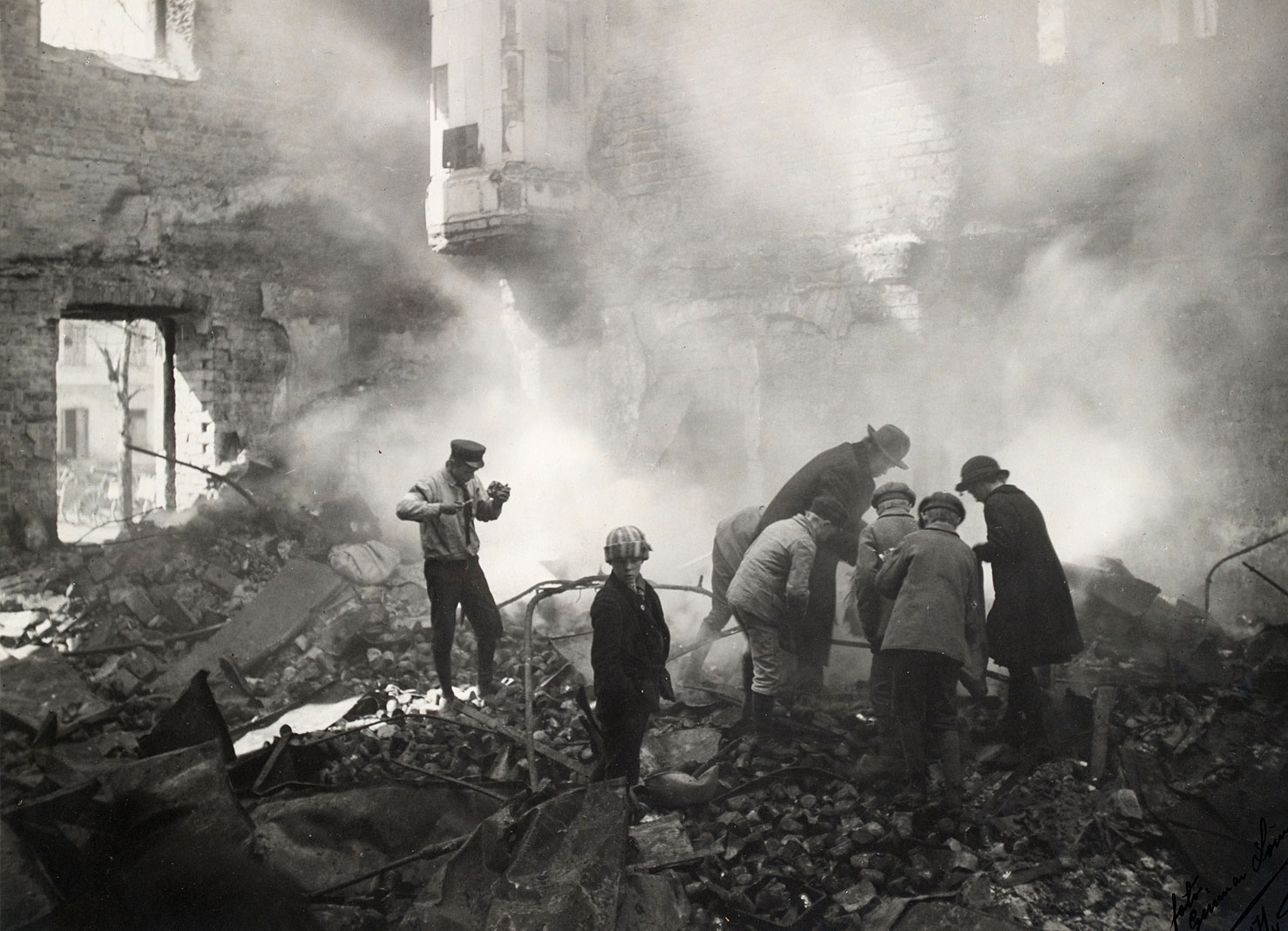 Valokuvassa tuhoutuneen rakennuksen jäännöksiä tutkimassa kaksi aikuista ja joukko lapsia. Raunioista nousee savu ja taustalla pystyssä säilyneitä tiiliseinän osia.