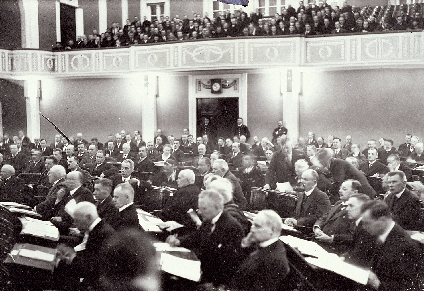 Valokuvassa kansanedustajia Heimolan salissa. Lehterillä yleisöä istuntoa kuuntelemassa.