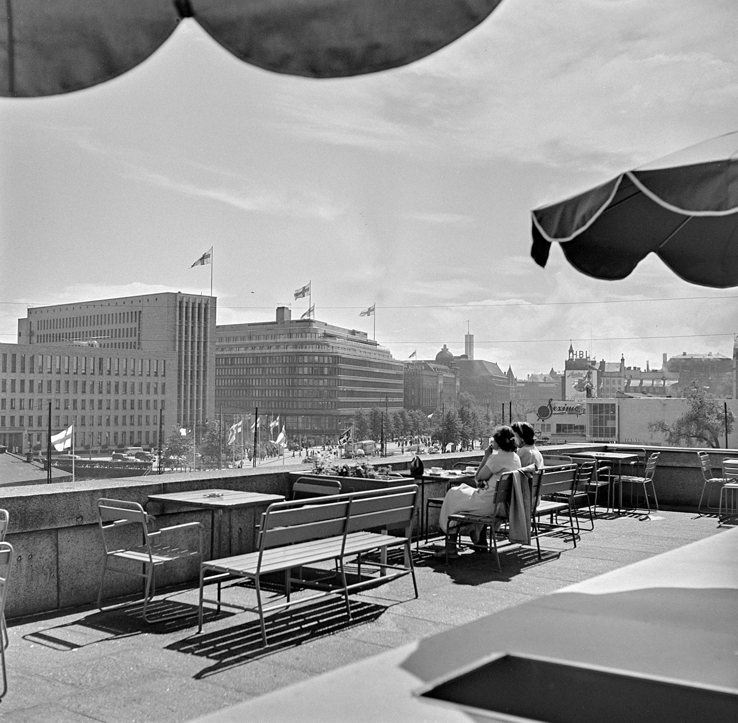 Valokuvassa näkymä eduskuntatalon katolta Postitalon ja Lasipalatsin suuntaan, jonne oli avattu olympialaisvieraiden kunniaksi terassi kesällä 1952. Kaksi naista istuu terassin pöydän ääressä selin kuvaajaan nähden.
